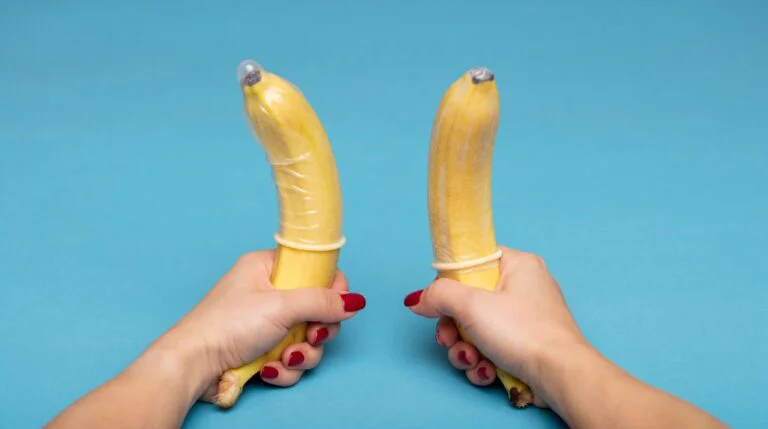 Hands with red fingernails holding two bananas wearing condoms against a blue background as an illustration of double penetration.